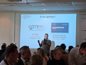 Dejeuner CPME 91 avec présentation sur l'écran en arrière plan, 1 homme avec micro et audience est autour de table de déjeuner.