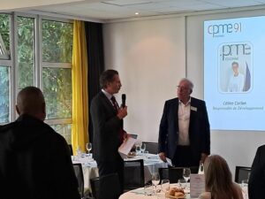Dejeuner CPME 91 avec présentation sur l'écran en arrière plan, 1 homme avec micro et 1 autre homme debout à coté. L'audience est autour de table de déjeuner.