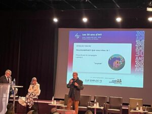 Scène des 30 de CAP Emploi 78 - IHY au Campus Oxygène Factory – Les Mureaux. 1 homme debout au pupitre, 1 femme assise, 1 homme debout au micro, présentation projetée en arrière plan sur l'écran