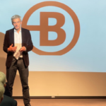 Intervention de Nicolas Potier au Café Rencontres de l'ADEZAC. Monsieur Potier sur scène, debout. En arrière plan logo Bruneau diffusé sur écran projecteur.